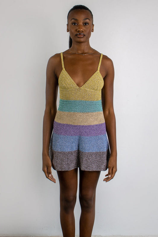 A woman stands against a plain white wall wearing a colorful striped knit romper with thin straps and a V-neckline. The romper features horizontal stripes in yellow, blue, purple, and gray-brown tones.